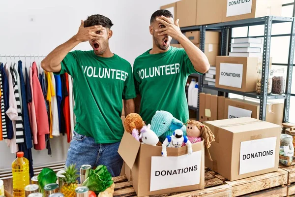 Young gay couple wearing volunteer t shirt at donations stand peeking in shock covering face and eyes with hand, looking through fingers with embarrassed expression.