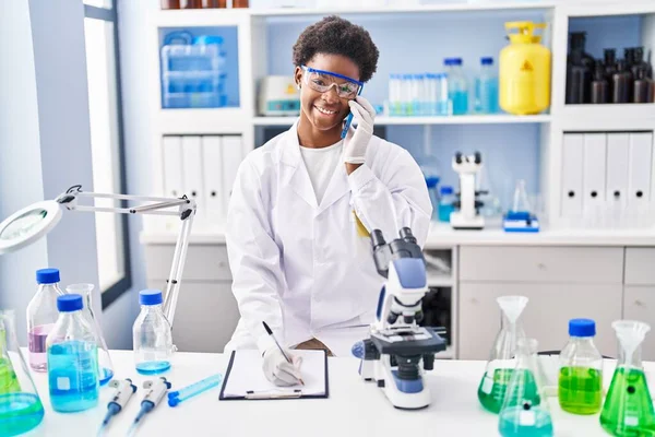 アフリカ系アメリカ人女性科学者の制服を着て研究室でクリップボードにスマートフォンの書き込みで話す — ストック写真