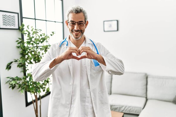 Middle age hispanic man wearing doctor uniform and stethoscope at waiting room smiling in love doing heart symbol shape with hands. romantic concept. 