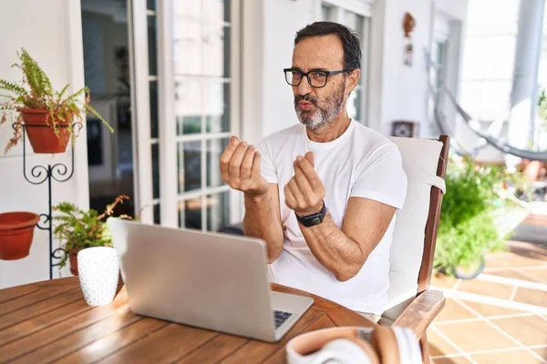 Middle age man using computer laptop at home doing money gesture with hands, asking for salary payment, millionaire business