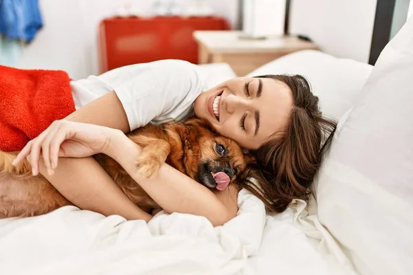 Young hispanic woman hugging dog lying on bed at bedroom