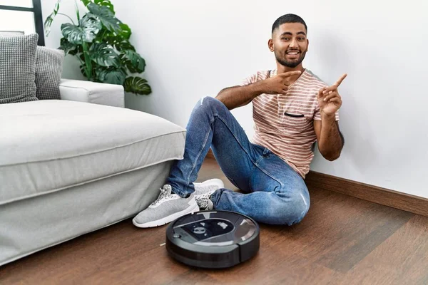 Young indian man sitting at home by vacuum robot smiling and looking at the camera pointing with two hands and fingers to the side.