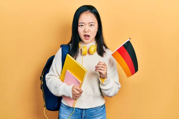 Young chinese girl exchange student holding germany flag in shock face, looking skeptical and sarcastic, surprised with open mouth