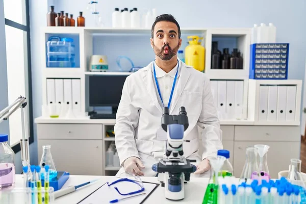 Young hispanic man with beard working at scientist laboratory making fish face with lips, crazy and comical gesture. funny expression.