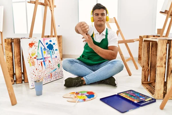 Young hispanic man at art studio looking unhappy and angry showing rejection and negative with thumbs down gesture. bad expression.