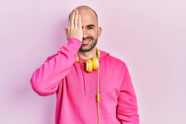 Spor kıyafetleri giyen ve kulaklık takan genç kel bir adam bir gözünü eliyle kapatıyor, yüzünde kendine güvenen bir gülümseme ve şaşırtıcı bir duygu var.. 