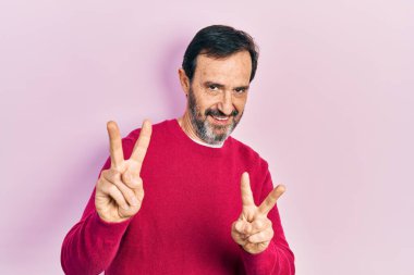 Orta yaşlı, sıradan kıyafetler giyen, gülümseyen, parmaklarını zafer işareti yaparken gösteren İspanyol bir adam. İki numara.. 