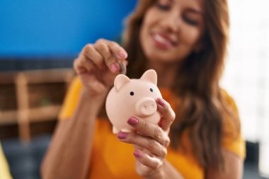 Genç Latin kadın gülümsüyor. Kendine güveni tam. Evdeki kumbaraya para koyuyor.