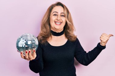 Orta yaşlı beyaz kadın, elinde parlak disko topu tutarak baş parmağını yana uzatıyor ve ağzı açık gülümsüyor. 