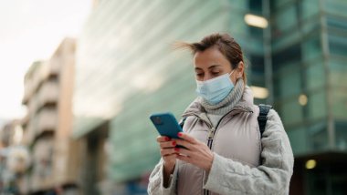 Orta yaşlı, tıbbi maske takan bir kadın sokakta akıllı telefon kullanıyor.