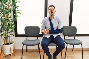 Yakışıklı genç adam, kolundan yaralanmış bir şekilde bekleme odasında oturuyor sinirli ve sinirli bir şekilde bağırıyor, öfkeyle bağırıyor. öfke ve agresif konsept. 