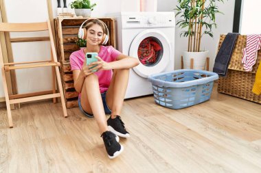 Çamaşır odasında çamaşır makinesi bekleyen akıllı telefon ve kulaklık kullanan beyaz bir kadın.
