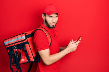 Hispanic man with beard wearing delivery package backpack using smartphone skeptic and nervous, frowning upset because of problem. negative person. 