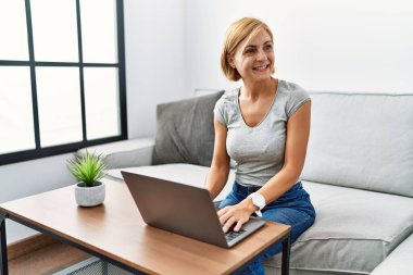 Orta yaşlı sarışın kadın evde dizüstü bilgisayar kullanıyor. Yüzünde gülümseme, doğal bir ifade ile yan yana bakıyor. Kendinden emin gülen. 