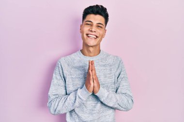 Gündelik kıyafetler giyen genç İspanyol adam elleriyle dua ediyor af diliyor kendinden emin bir şekilde gülümsüyor.. 