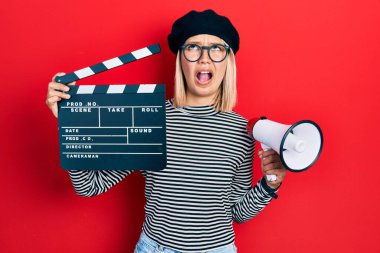 Beautiful blonde woman holding video film clapboard and megaphone angry and mad screaming frustrated and furious, shouting with anger looking up. 