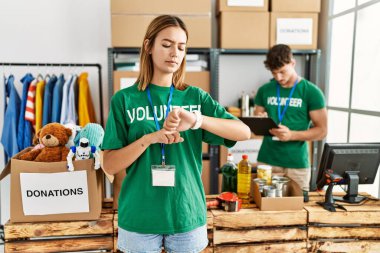 Young blonde girl wearing volunteer t shirt at donation stand checking the time on wrist watch, relaxed and confident 