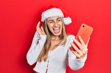 Güzel İspanyol kadın Noel şapkası takıyor. Akıllı telefon kullanıyor. Çılgınlığı kutluyor ve başarıyı hayrete düşürüyor.. 