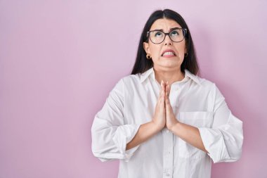 Genç esmer kadın pembe arka planda duruyor. Elleriyle yalvarıyor ve dua ediyor. Yüzünde umut ifadesi var. Yalvarıyor. 