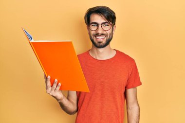 Young hispanic man reading book looking positive and happy standing and smiling with a confident smile showing teeth 