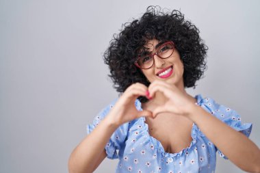 Kıvırcık saçlı, gözlüklü, izole edilmiş arka planda gülümseyen, elleriyle kalp sembolü yapan genç esmer kadın. romantik kavram. 