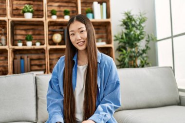 Genç Çinli kız gülümsüyor. Evdeki kanepede mutlu mesut oturuyor..