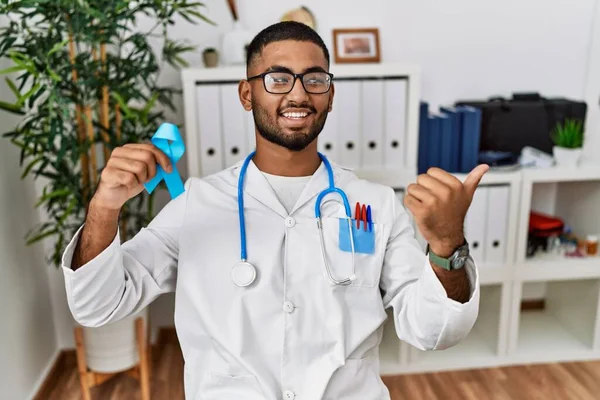 Young indian doctor holding blue ribbon pointing thumb up to the side smiling happy with open mouth
