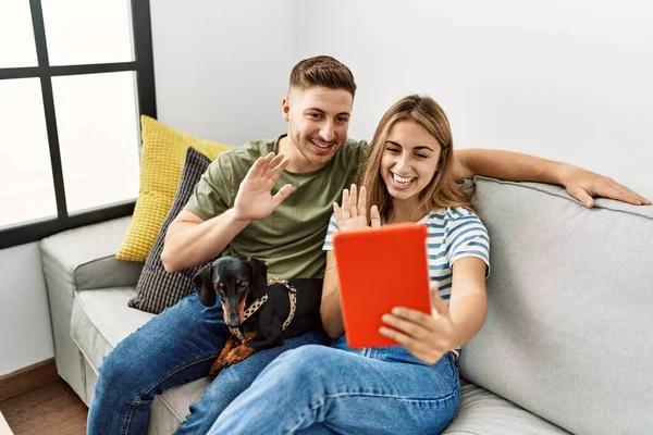 Young hispanic couple having video call using touchpad sitting on the sofa with dog at home.