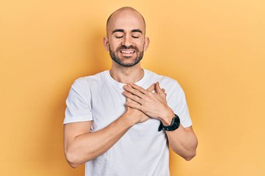 Gündelik beyaz tişört giyen genç kel adam elleri göğsünde, gözleri kapalı ve yüzünde minnet dolu bir jest ile gülümsüyor. sağlık kavramı. 
