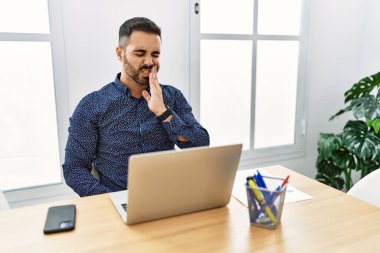 Ofiste sakallı, laptopuyla çalışan, diş ağrısı ya da diş hastalığından dolayı acı dolu bir ifadeyle dudaklarına dokunan genç İspanyol bir adam. dişçi 