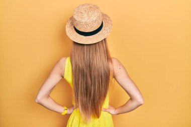 Sarışın, yaz şapkası takan genç kız kolları vücudunda geriye doğru bakıyor. 