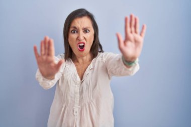 Middle age hispanic woman standing over blue background doing stop gesture with hands palms, angry and frustration expression 