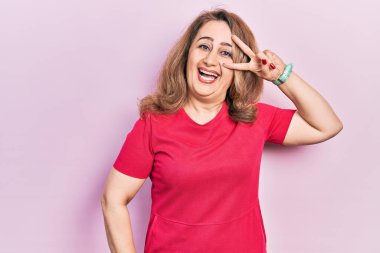 Orta yaşlı beyaz kadın, günlük kıyafetler giyip barış sembolü çiziyor. Yüzünde parmaklar, gülümseyerek zaferi gösteriyor. 