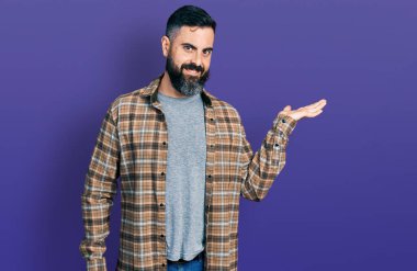 Sakallı, sıradan tişörtlü, neşeli gülümseyen, elini kaldırıp kameraya bakan İspanyol bir adam.. 