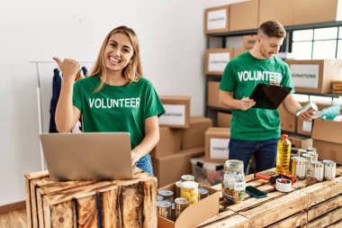 Young woman and man wearing volunteer t shirt at donations stand pointing thumb up to the side smiling happy with open mouth 