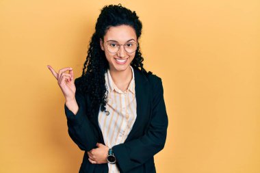 Young hispanic woman with curly hair wearing business clothes smiling happy pointing with hand and finger to the side 
