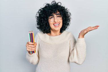 Orta Doğulu genç bir kadın elinde renkli kalemlerle başarıyı mutlu bir gülümsemeyle kutluyor ve kazanan el kaldırarak ifade ediyor. 