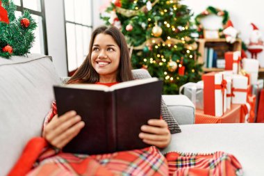 Genç Latin kadın kitap okuyor. Yılbaşı ağacının yanında oturuyor.