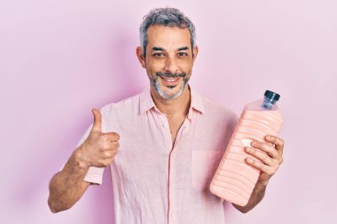Gri saçlı, elinde deterjan şişesi olan yakışıklı orta yaşlı bir adam mutlu ve pozitif gülümsüyor, başparmağıyla mükemmel ve onay işareti yapıyor. 