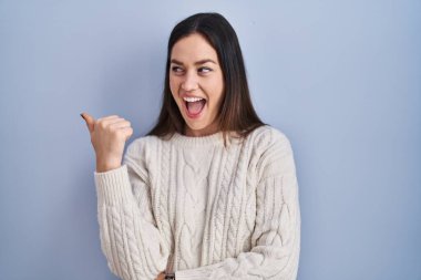 Mavi arka planda dikilen esmer kadın gülümsüyor, gülümsüyor ve başparmağıyla yan tarafa bakıyor.. 