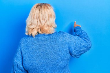 Middle age blonde woman wearing casual clothes posing backwards pointing ahead with finger hand 
