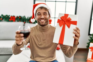 Genç İspanyol adam gülümsüyor, kendine güveniyor, elinde Noel hediyesi tutuyor ve evde şarap içiyor.