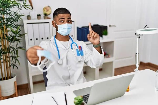 Young indian doctor offering safety mask surprised with an idea or question pointing finger with happy face, number one