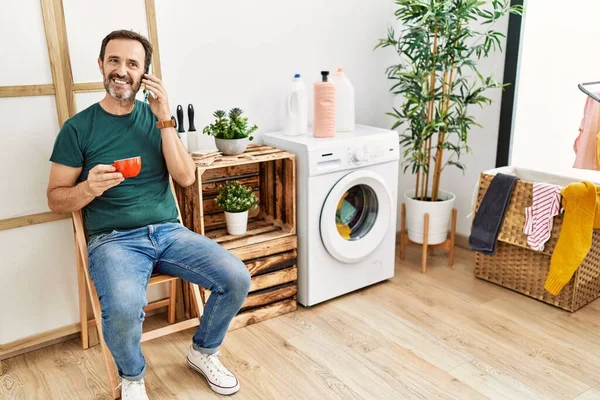 Middle Age Hispanic Man Doing Laundry Talking Smartphone Drinking Coffee — стоковое фото