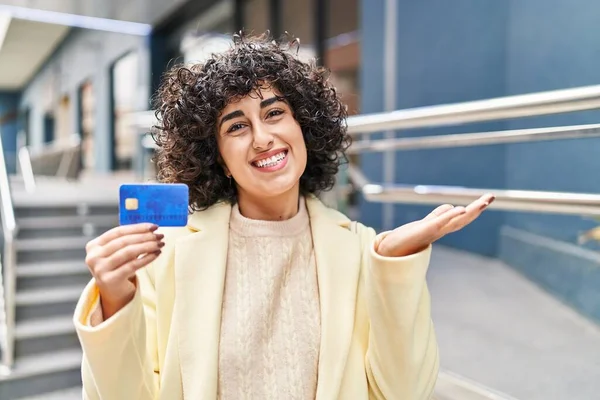 Young brunette woman with curly hair holding credit card celebrating achievement with happy smile and winner expression with raised hand