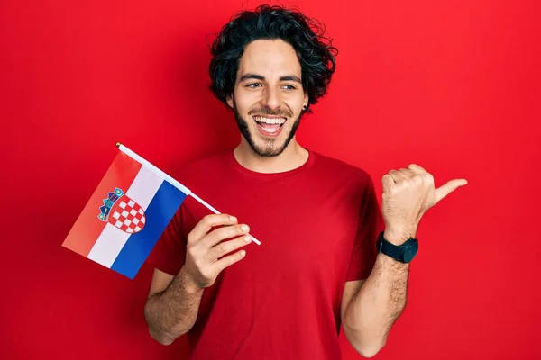 英俊的他的惊慌失措的男人举着克罗地亚国旗 把大拇指指向侧面 笑容满面 张开嘴 — 图库照片