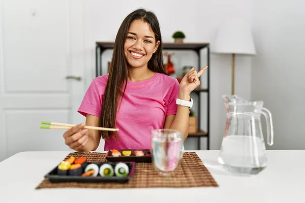 若いブルネットの女性がカメラを見ている側に手指で指して 顔に大きな笑顔で箸を使って寿司を食べる — ストック写真