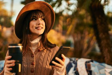 Kışlık şapkalı esmer kadın akıllı telefon kullanıyor ve parkta kahve içiyor.