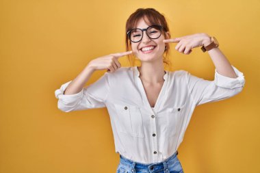 Sarı arka plan üzerine gündelik tişört giyen, neşeli gülümseyen, parmaklarını ve ağzını gösteren genç ve güzel bir kadın. diş sağlığı kavramı. 