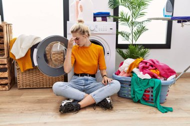 Young blonde woman doing laundry sitting by washing machine tired rubbing nose and eyes feeling fatigue and headache. stress and frustration concept. 